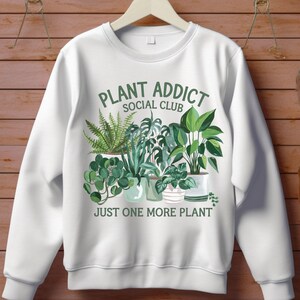 May include: White sweatshirt with a green and white graphic of potted plants. The text "Plant Addict Social Club" and "Just One More Plant" is printed on the sweatshirt.