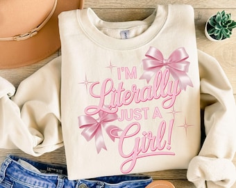 May include: A cream colored sweatshirt with the text "I'm Literally Just a Girl!" in pink script with pink bows.