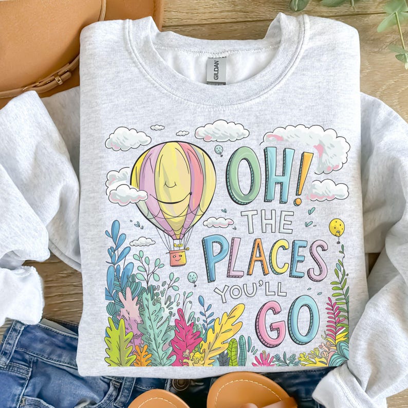 May include: A grey sweatshirt featuring a colourful hot air balloon, clouds and plants. The text "Oh! The Places You'll Go" is printed on the sweatshirt.