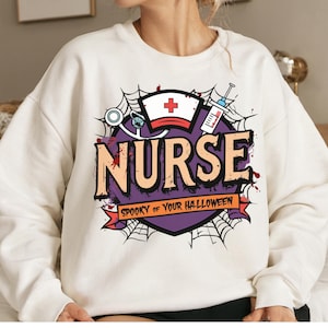 Op de afbeelding: Crèmekleurige sweatshirt met een Halloween-thema. Het ontwerp bevat het woord "NURSE" in oranje en zwart, een verpleegstersmuts, een spuit en de zin "SPOOKY OF YOUR HALLOWEEN". Spinnenwebben en bloedspetters maken het ontwerp compleet.