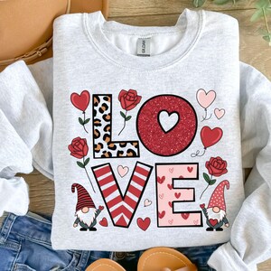 Op de afbeelding: Een grijs sweatshirt met een Valentijnsdagontwerp met het woord "LOVE" in kleurrijke letters met harten, gnomen en bloemen.
