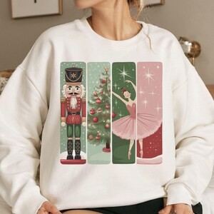 Op de afbeelding: Een witte sweatshirt met een feestelijk ontwerp. Het ontwerp toont vier verticale panelen: een notenkraker, een kerstboom en een ballerina in een roze tutu. Het ontwerp heeft een vakantiethema met een vleugje ballet.