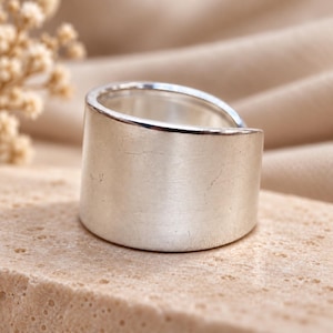 Peut inclure: Une large bague anneau en argent poli avec une surface lisse et continue et un bord supérieur légèrement incliné. Le métal présente une finition réfléchissante avec de fines marques de surface subtiles visibles sur l'extérieur de l'anneau.
