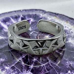 Könnte beinhalten: Verstellbarer Ring in Silber mit einem geometrischen Sternmuster. Der Ring ist innen mit "S925" gestempelt. Das Design des Rings zeigt eine Reihe erhabener Sterne, die eine strukturierte Oberfläche bilden. Der Ring wird auf einer lila-weißen Oberfläche präsentiert.