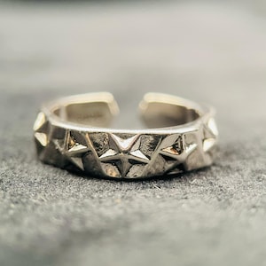 Op de afbeelding: Een zilverkleurige metalen ring met een open bandontwerp. Het buitenoppervlak van de ring is versierd met een herhalend patroon van verhoogde, vijfpuntige sterren die een textuurrijke, licht donkere afwerking hebben tussen de punten.