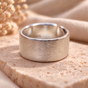 Op de afbeelding: Een brede zilverkleurige ring met een geborstelde, matte afwerking aan de buitenzijde. De ring heeft een open ontwerp met een zichtbare opening aan de achterkant, en aan de binnenzijde is een klein gestempeld keurmerk bij de opening te zien.