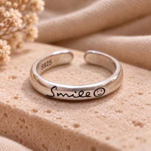 Peut inclure: Une bague ouverte de couleur argentée portant le mot Smile écrit en lettres cursives noires, suivi d'une petite icône de visage souriant. L'intérieur de l'anneau est clairement marqué de l'inscription S925, indiquant la composition du métal.