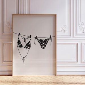 Könnte beinhalten: Ein gerahmter Druck mit einer schwarz-weißen Illustration eines Bikinis. Das Ober- und Unterteil des Bikinis hängen mit Wäscheklammern an einer Wäscheleine. Der Bikini hat ein gepunktetes Muster. Der Rahmen ist hellbraun.