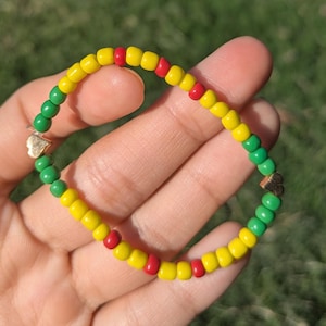 May include: A beaded bracelet featuring yellow, red, and green beads. The bracelet has a Rastafarian color scheme, with gold-colored accents. The bracelet is held in a hand against a blurred green background.