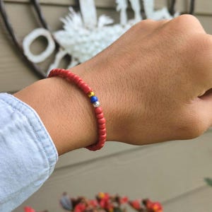 May include: Red beaded bracelet with gold and blue accent beads. The bracelet is worn on a wrist, with a denim shirt sleeve visible in the background. The bracelet is a simple, yet colorful accessory.
