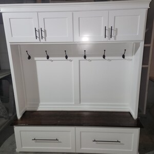 May include: White wooden entryway bench with two drawers and a top shelf with three cabinets. The bench has a dark wood top and black metal hooks.