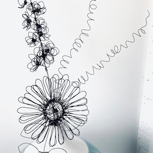 May include: A black wire sculpture of flowers in a blue and white ceramic vase. The flowers are made of wire and are arranged in a bouquet. The vase is a simple, round shape with a blue glaze.