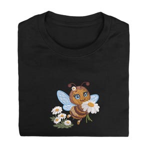 May include: A black t-shirt featuring an embroidered design of a brown bee with blue wings, holding a white daisy. The bee has large blue eyes and a small white flower on its head. Below the bee are three additional white daisies with green stems.
