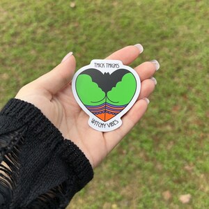 May include: A heart-shaped sticker with a green and black design, featuring the words "THICK THIGHS" and "WATCHY VIBES". The sticker is held in a hand against a blurred green background.