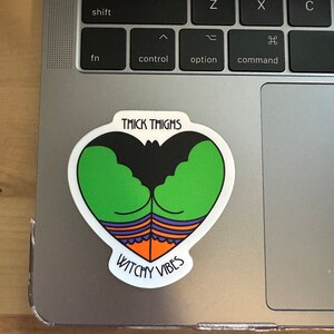 May include: A heart-shaped sticker with a cartoon design featuring green thighs, a black bat-like shape, and orange and purple accents. The sticker has the text "THICK THIGHS" at the top and "WITCHY VIBES" at the bottom.
