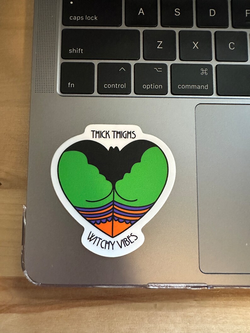 May include: A heart-shaped sticker with a cartoon design featuring green thighs, a black bat-like shape, and orange and purple accents. The sticker has the text "THICK THIGHS" at the top and "WITCHY VIBES" at the bottom.