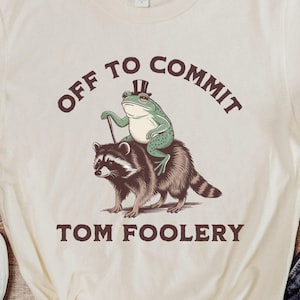 Puede incluir: Una camiseta blanca con un gráfico de una rana con un sombrero de copa montando un mapache. El texto "Off to commit tom foolery" está impreso en la camiseta.