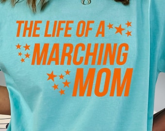 Może przedstawiać: Jasnoniebieska koszulka z pomarańczowym napisem "THE LIFE OF A MARCHING MOM" i gwiazdkami. Koszulka z krótkim rękawem ma luźny krój, idealna na co dzień.
