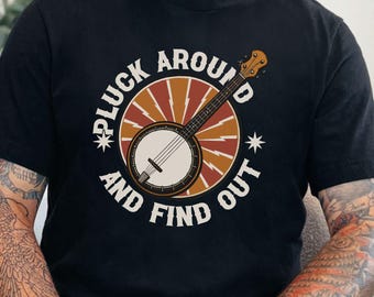 Könnte beinhalten: Schwarzes T-Shirt mit einem kreisförmigen Design, das ein Banjo und den Text "PLUCK AROUND AND FIND OUT" zeigt. Das Design hat orange, braune und weiße Elemente. Das T-Shirt hat einen Rundhalsausschnitt.