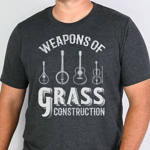Op de afbeelding: Een donkergrijs T-shirt met een witte afbeelding van muziekinstrumenten en de tekst "Weapons of Grass Construction".