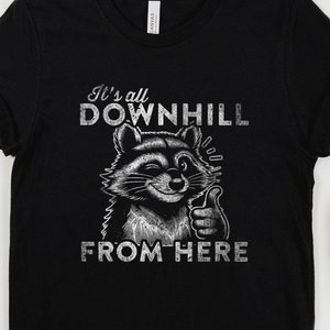 May include: Black t-shirt featuring a white graphic of a raccoon giving a thumbs up. The text on the shirt reads "It's all downhill from here."