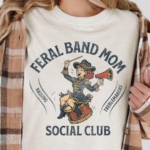 May include: Cream-colored t-shirt with a vintage-style graphic. The graphic features a cartoon band leader with a megaphone, drum, and the text "FERAL BAND MOM SOCIAL CLUB". The shirt is worn by a person with long blonde hair and a brown plaid shirt.