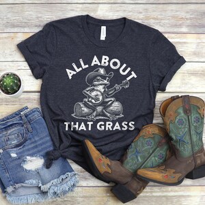 May include: Dark gray t-shirt with the text "All About That Grass" and a frog playing a banjo. The shirt is paired with denim shorts and brown cowboy boots with green and blue accents.