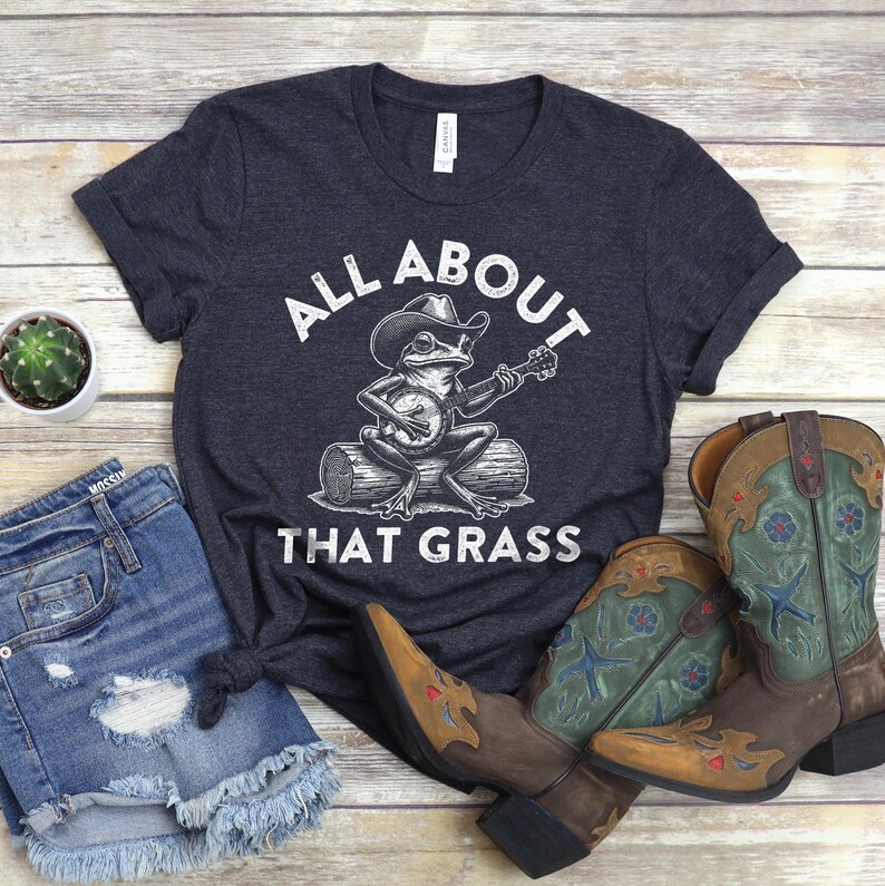 May include: Dark gray t-shirt with the text "All About That Grass" and a frog playing a banjo. The shirt is paired with denim shorts and brown cowboy boots with green and blue accents.