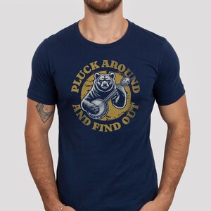Pu&ograve; includere: Una t-shirt blu navy con una grafica di un orso che suona il banjo. La grafica &egrave; circondata da un cerchio con il testo "Pluck Around And Find Out".