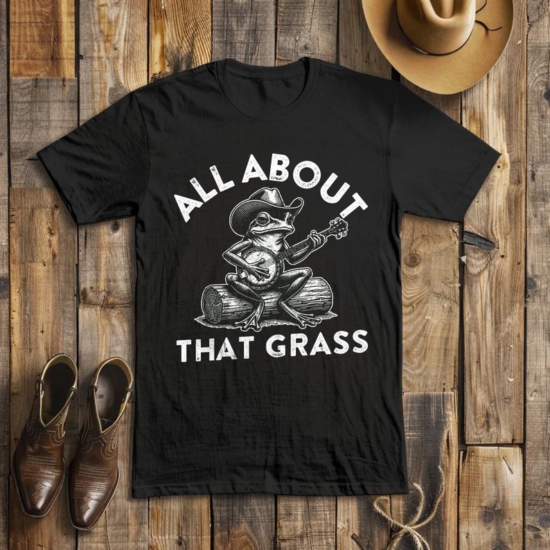 May include: Black t-shirt with a graphic of a frog wearing a cowboy hat and playing a banjo on a log. The text "ALL ABOUT THAT GRASS" is printed above and below the frog illustration.