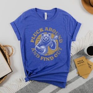 Pu&ograve; includere: Una t-shirt blu reale con una grafica di un orso che suona il banjo. La grafica &egrave; circondata dal testo "Pluck Around And Find Out".
