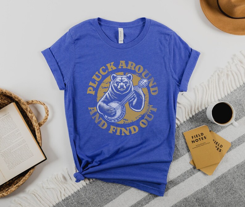 Pu&ograve; includere: Una t-shirt blu reale con una grafica di un orso che suona il banjo. La grafica &egrave; circondata dal testo "Pluck Around And Find Out".