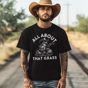 May include: Black t-shirt with the text "ALL ABOUT THAT GRASS" and a graphic of a frog playing a banjo while wearing a cowboy hat. The frog is sitting on a log. The person in the image is wearing a brown cowboy hat.