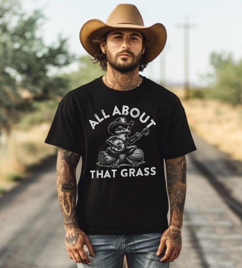 May include: Black t-shirt with the text "ALL ABOUT THAT GRASS" and a graphic of a frog playing a banjo while wearing a cowboy hat. The frog is sitting on a log. The person in the image is wearing a brown cowboy hat.
