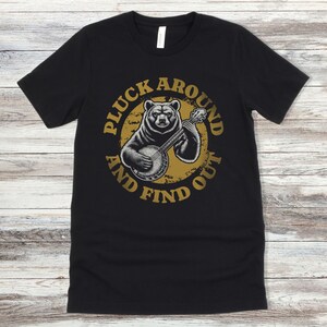 Pu&ograve; includere: Maglietta nera con un grafico circolare dorato invecchiato che presenta un orso che suona un banjo. Il testo "Pluck Around And Find Out" &egrave; scritto attorno al cerchio.