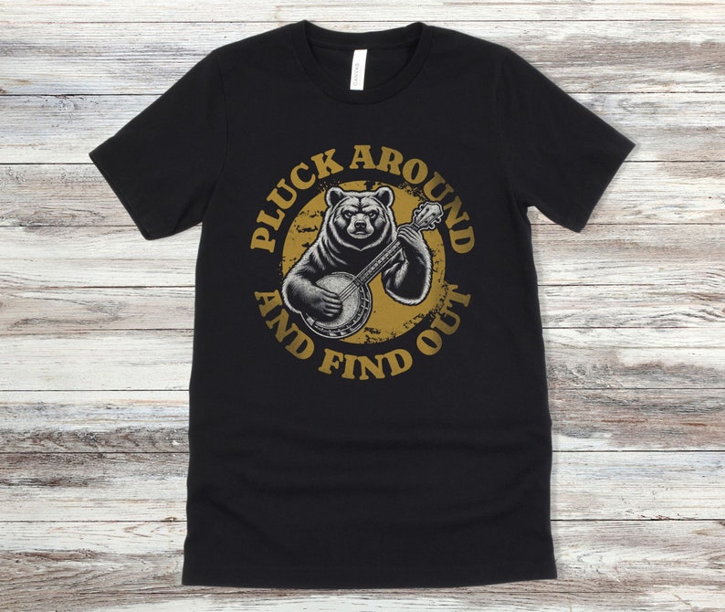 Pu&ograve; includere: Maglietta nera con un grafico circolare dorato invecchiato che presenta un orso che suona un banjo. Il testo "Pluck Around And Find Out" &egrave; scritto attorno al cerchio.