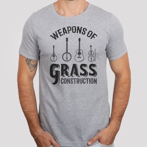 Op de afbeelding: Een grijs T-shirt met de tekst "Weapons of Grass Construction" in zwart. De tekst wordt omringd door een cirkel van vijf muziekinstrumenten: een banjo, een gitaar, een mandoline, een viool en een plectrum.