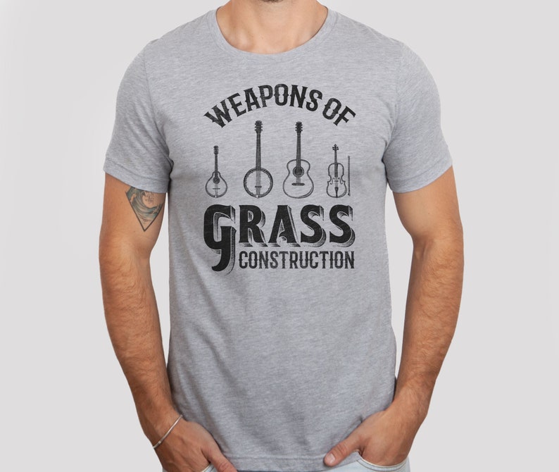 Op de afbeelding: Een grijs T-shirt met de tekst "Weapons of Grass Construction" in zwart. De tekst wordt omringd door een cirkel van vijf muziekinstrumenten: een banjo, een gitaar, een mandoline, een viool en een plectrum.