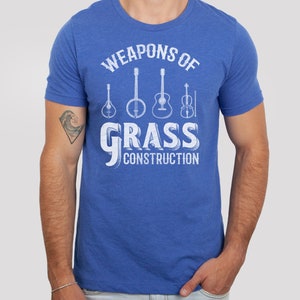 Op de afbeelding: Een koninklijk blauw t-shirt met een witte afbeelding van muziekinstrumenten en de tekst "Weapons of Grass Construction".