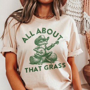 May include: Beige t-shirt with the text "ALL ABOUT THAT GRASS" in green. The shirt features a green illustration of a frog wearing a cowboy hat and playing a banjo while sitting on a log.