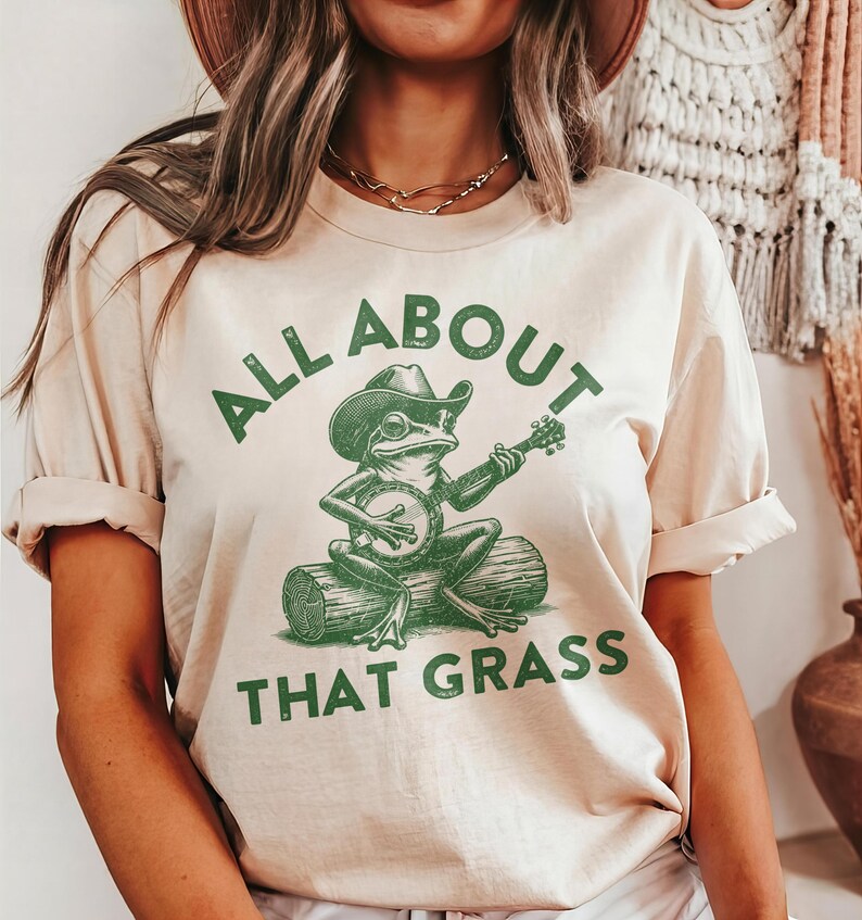 May include: Beige t-shirt with the text "ALL ABOUT THAT GRASS" in green. The shirt features a green illustration of a frog wearing a cowboy hat and playing a banjo while sitting on a log.