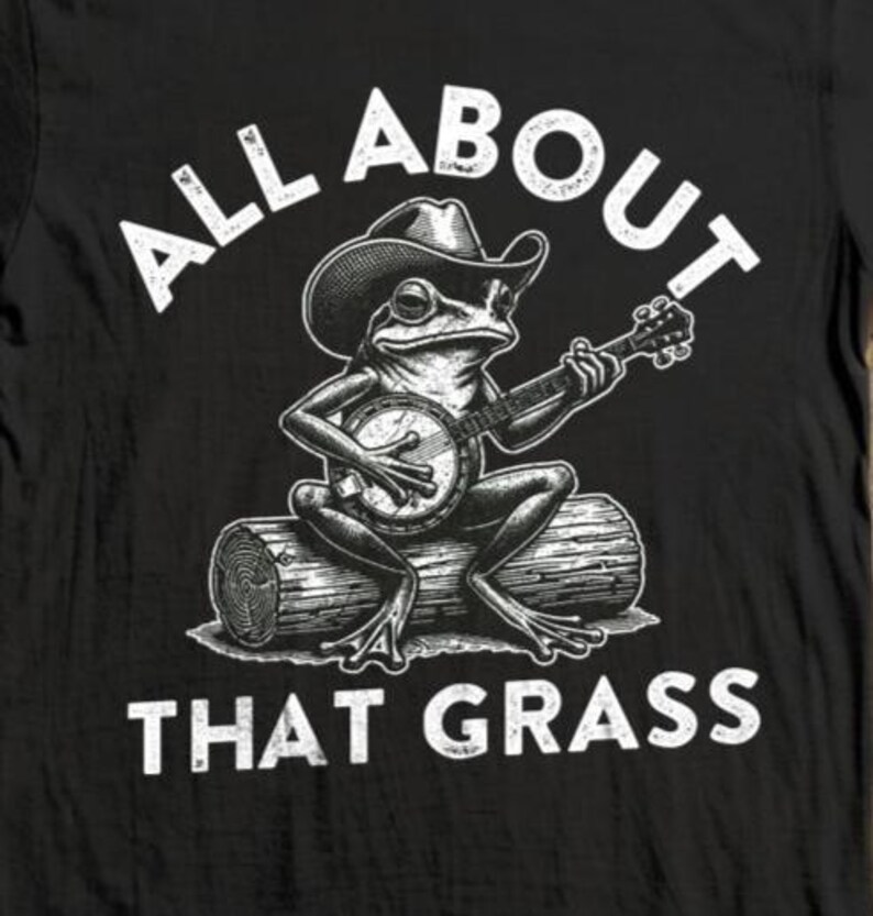 May include: Black t-shirt featuring a frog wearing a cowboy hat and playing a banjo while sitting on a log. The text "ALL ABOUT THAT GRASS" is printed above and below the frog illustration in white.