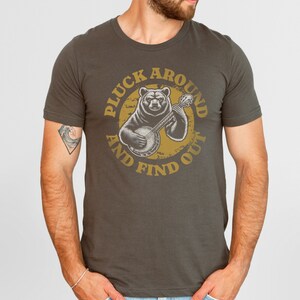 Pu&ograve; includere: Una maglietta grigio scuro con una grafica di un orso che suona il banjo. La grafica &egrave; in un cerchio con il testo "Pluck Around And Find Out" in un font giallo.