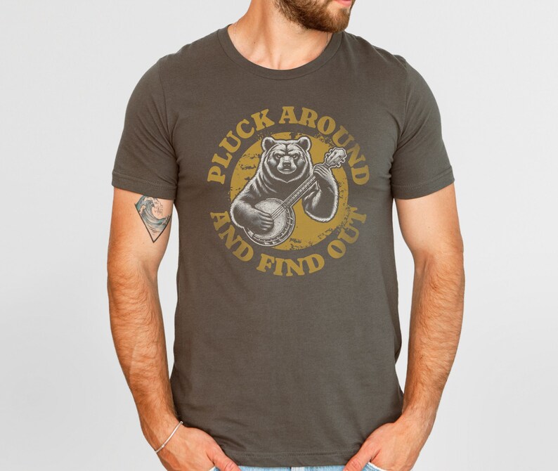 Pu&ograve; includere: Una maglietta grigio scuro con una grafica di un orso che suona il banjo. La grafica &egrave; in un cerchio con il testo "Pluck Around And Find Out" in un font giallo.