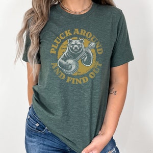 Pu&ograve; includere: Una maglietta verde con un'immagine di un orso che suona il banjo all'interno di un cerchio con il testo "Pluck Around And Find Out".