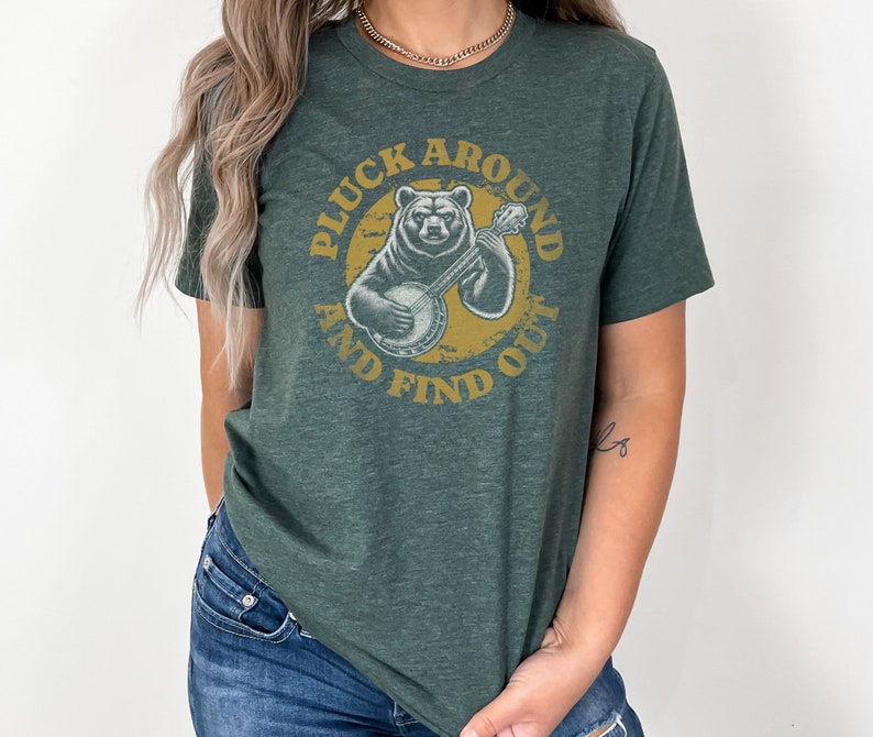 Pu&ograve; includere: Una maglietta verde con un'immagine di un orso che suona il banjo all'interno di un cerchio con il testo "Pluck Around And Find Out".