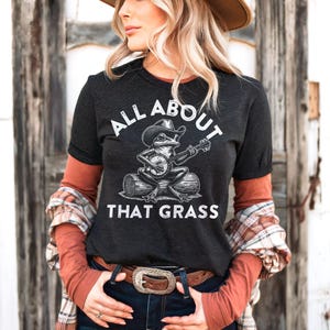 May include: Dark gray t-shirt with the text "ALL ABOUT THAT GRASS" and a graphic of a frog playing a banjo while wearing a cowboy hat. The shirt is paired with a long-sleeved orange shirt and a plaid shirt.
