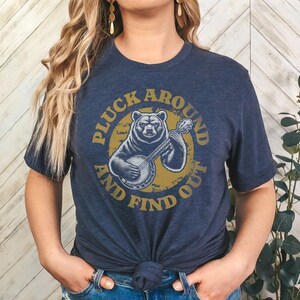 Pu&ograve; includere: Una maglietta blu navy con una grafica di un orso che suona il banjo. La grafica &egrave; in un cerchio con il testo "Pluck Around And Find Out" in un font giallo.