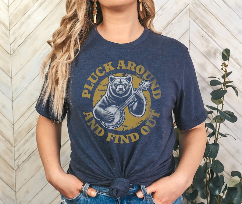 Pu&ograve; includere: Una maglietta blu navy con una grafica di un orso che suona il banjo. La grafica &egrave; in un cerchio con il testo "Pluck Around And Find Out" in un font giallo.