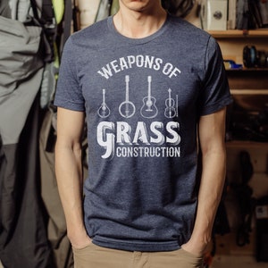 Op de afbeelding: Een marineblauw t-shirt met een wit grafisch ontwerp met de tekst "Weapons of Grass Construction" en een rij van vijf muziekinstrumenten, waaronder een banjo, een gitaar en een viool.